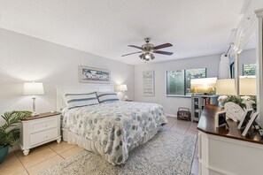 Une chambre à coucher comprenant un lit, des tables de chevet, un ventilateur de plafond, une fenêtre avec des stores, une bibliothèque et un miroir.
