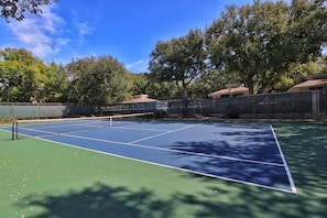 En udendørs tennisbane med net, omgivet af træer og et hegn.