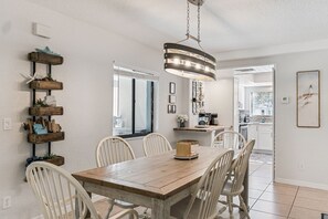 Un comedor con una mesa de madera y sillas blancas, un lámpara colgante, y una cocina visible a través de una puerta abierta.