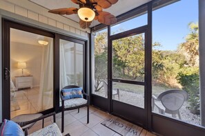 Un patio cubierto con ventilador de techo, una silla de mimbre con cojín y vistas a un jardín a través de amplias puertas de vidrio.