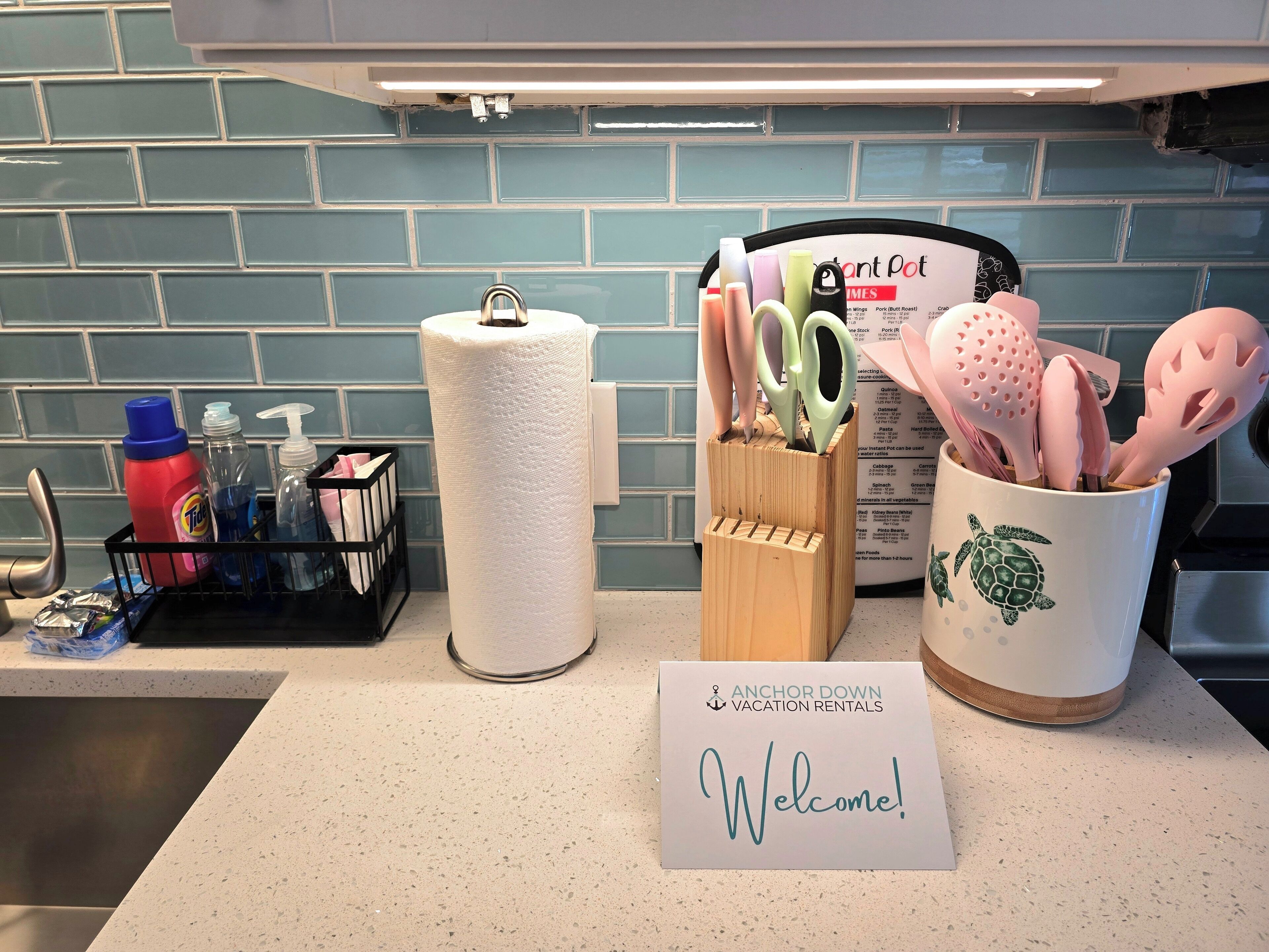 Eine Küchenarbeitsplatte mit einem Willkommensschild, einem Papiertuchhalter, einem Geschirrgestell mit Reinigungsmitteln und einer Tasse mit Küchenutensilien.