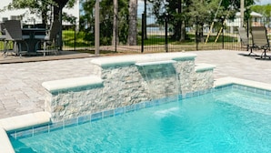 A swimming pool with a stone waterfall feature, surrounded by a paved patio with outdoor furniture and a fence.