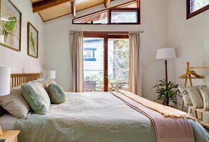 A bedroom with a bed, a nightstand, a lamp, a chair, a plant, and a window with curtains. - Dove Studio with Garden Patio (Byron Bay)