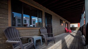 Een houten veranda met Adirondack-stoelen en een kleine tafel, een rode stoel en een raam dat een gebouw weerspiegelt.