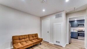 Een kamer met een bruine bank, witte muren en een keuken die door een open deur zichtbaar is.
