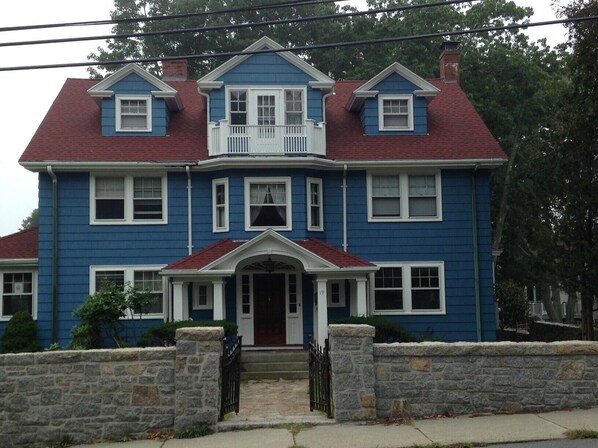 A two-story blue house with a red roof, white trim, and a stone wall in front.
