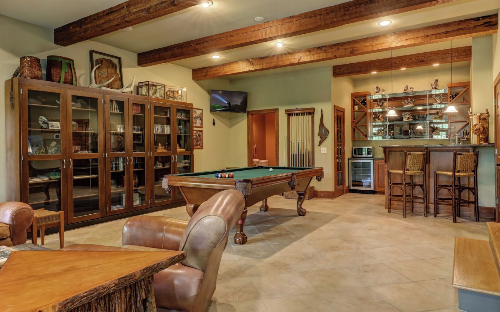 A spacious room with a pool table, leather chairs, a bar area, and a wooden cabinet with glass doors.
