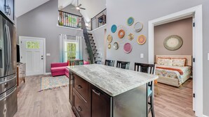 Una cocina amplia con una isla central, un comedor con sillas, una escalera y un dormitorio con una cama y detalles decorativos en la pared.