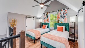 Un dormitorio con dos camas, un ventilador de techo, una ventana con cortinas florales y un estante pequeño.