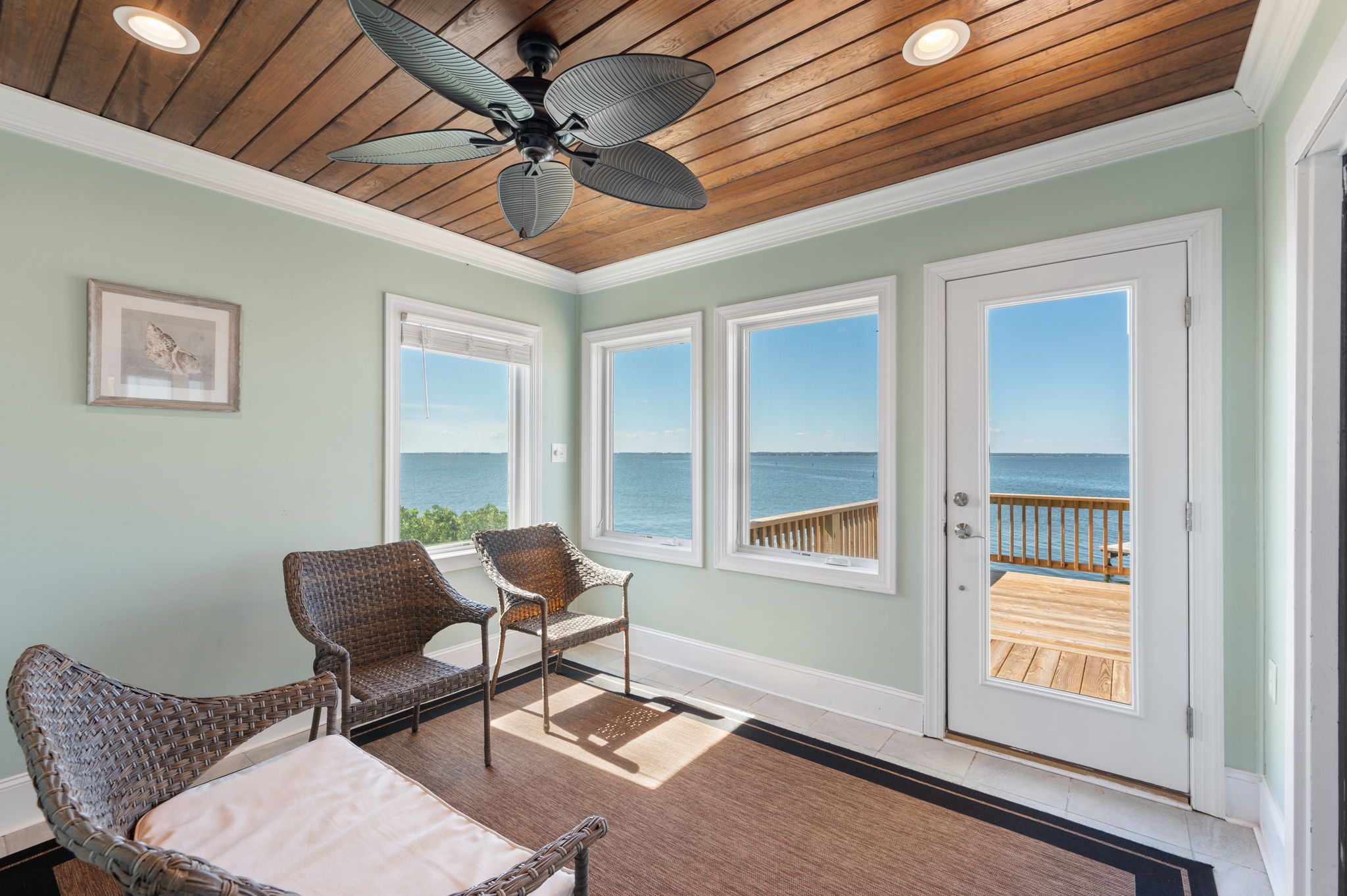 A room with a ceiling fan, large windows, wicker furniture, and a view of the sea.