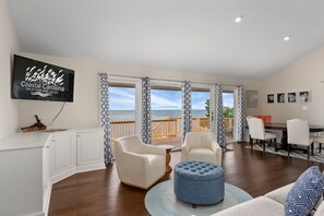 Un salon spacieux avec une grande télévision, deux fauteuils blancs, un pouf bleu et un coin repas comprenant une table et des chaises en bois.