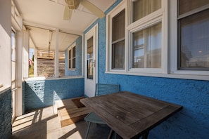 A covered outdoor area with a wooden table and chairs, a door, and windows with white frames.