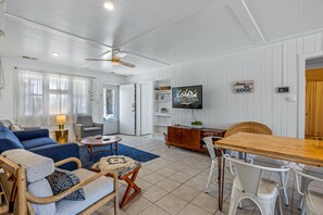 A spacious living room with a blue sofa, a wooden coffee table, and a dining table with white chairs. A television is mounted on the wall, and there is a ceiling fan.