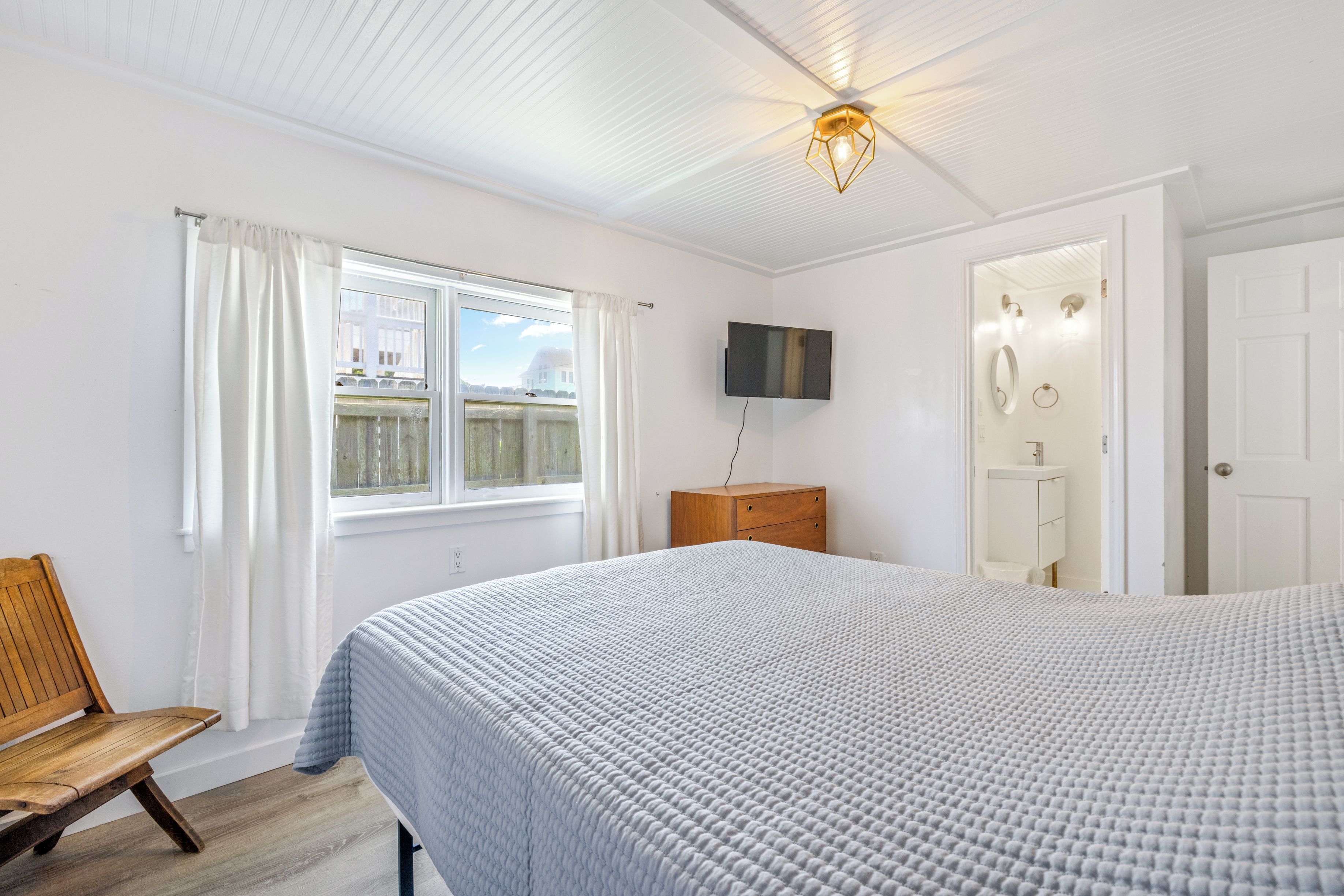 A bedroom with a large bed, a wooden bench, a TV mounted on the wall, and a window with curtains.