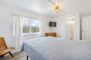A bedroom with a large bed, a wooden bench, a TV mounted on the wall, and a window with curtains.
