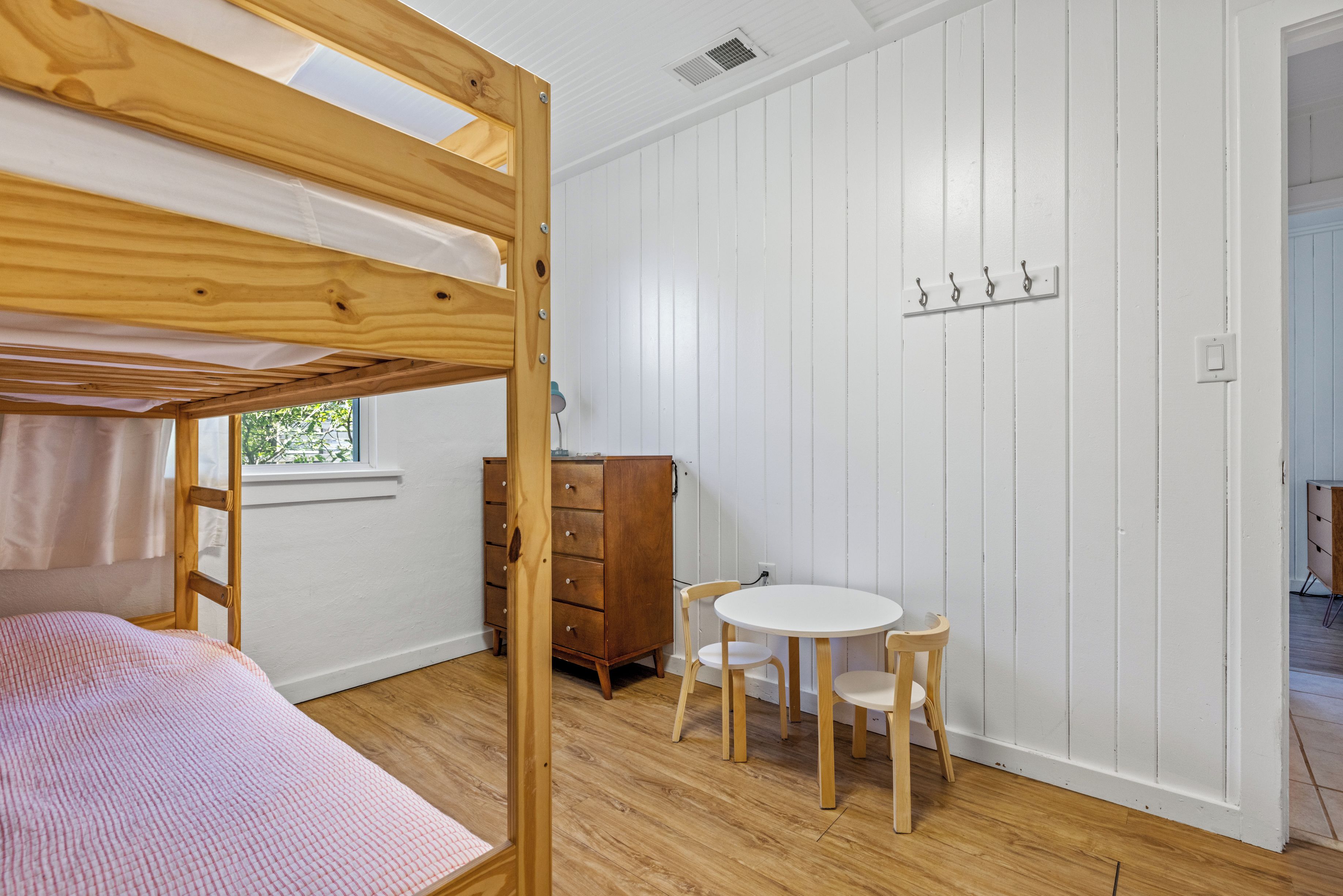 Una cama litera con un edredón rosa, un aparador de madera, una mesa redonda con sillas y una ventana que ofrece vistas al verdor.