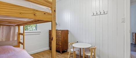 Un lit superposé avec une couverture rose, une commode en bois, une table ronde avec des chaises et une fenêtre donnant sur un paysage verdoyant.