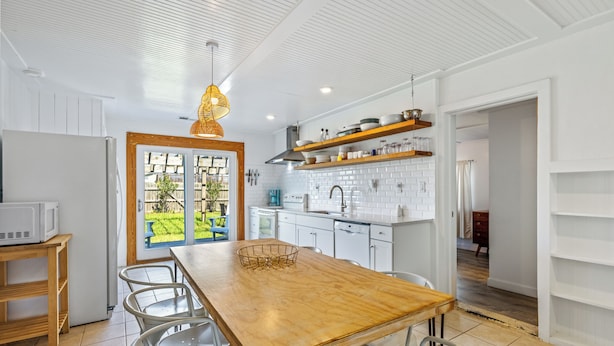 A modern kitchen with white cabinetry, a wooden dining table, and a view through a glass door to a garden. - Lunar Tide: Pet Friendly, Oceanside, Close to Beach and Avalon Pier (Kill Devil Hills)
