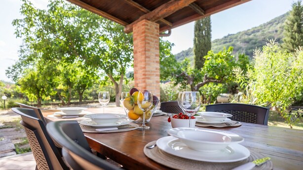 An outdoor dining table set with plates, glasses, and a bowl of fruit, surrounded by chairs and a brick pillar, with a view of trees and hills in the background. - Andrea and Carolina Domus (Castiglione Di Sicilia)