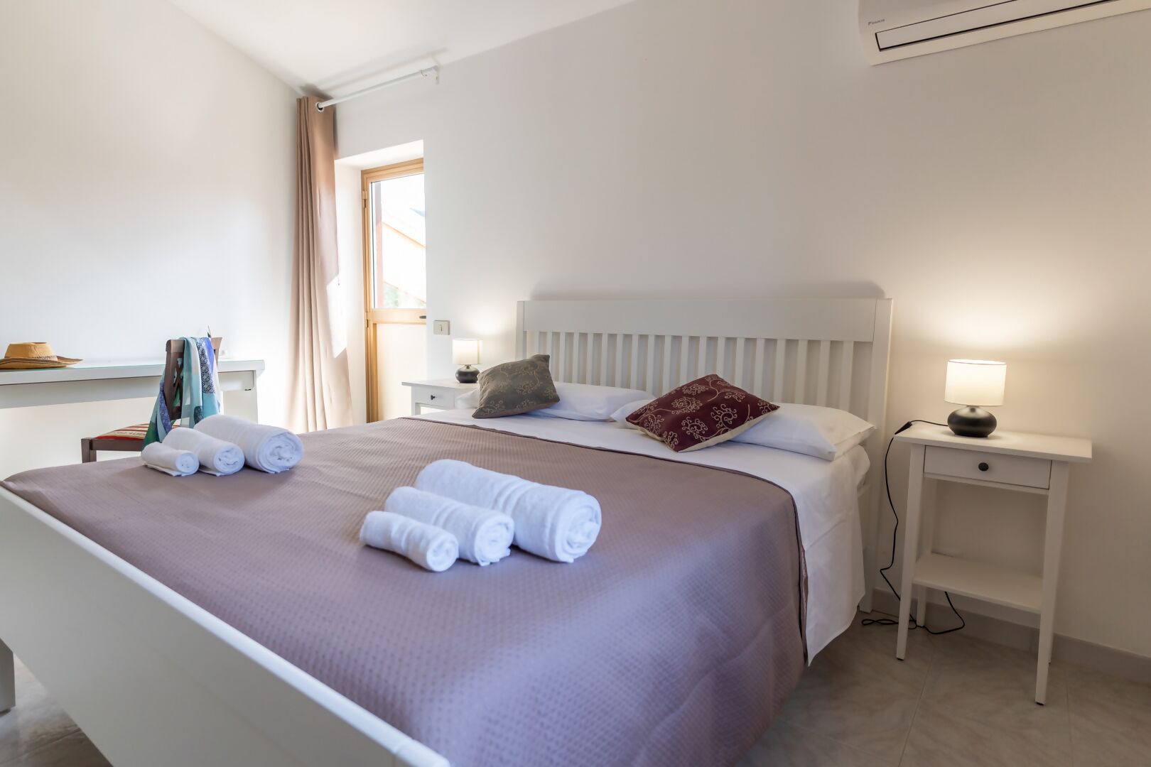 Una cama bien hecha con toallas enrolladas, una lámpara de noche y una ventana con cortinas.