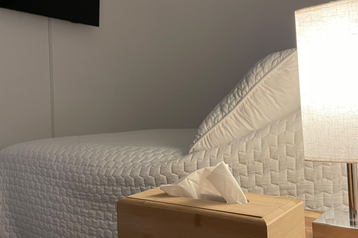 En pænt redt seng med et tykt hvidt dynebetræk, et trænatbord med en kasse toiletpapir og en lampe med hvid skærm.