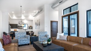 Ein modernes Wohnzimmer mit einer braunen Ledersofa, gemusterten Sesseln, einem dunklen Couchtisch und einem großen Fenster mit Jalousien.