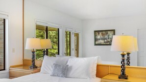 Quarto de hotel com uma cama grande, duas mesas de cabeceira com abajures, uma janela com vista para vegetação e um quadro na parede.