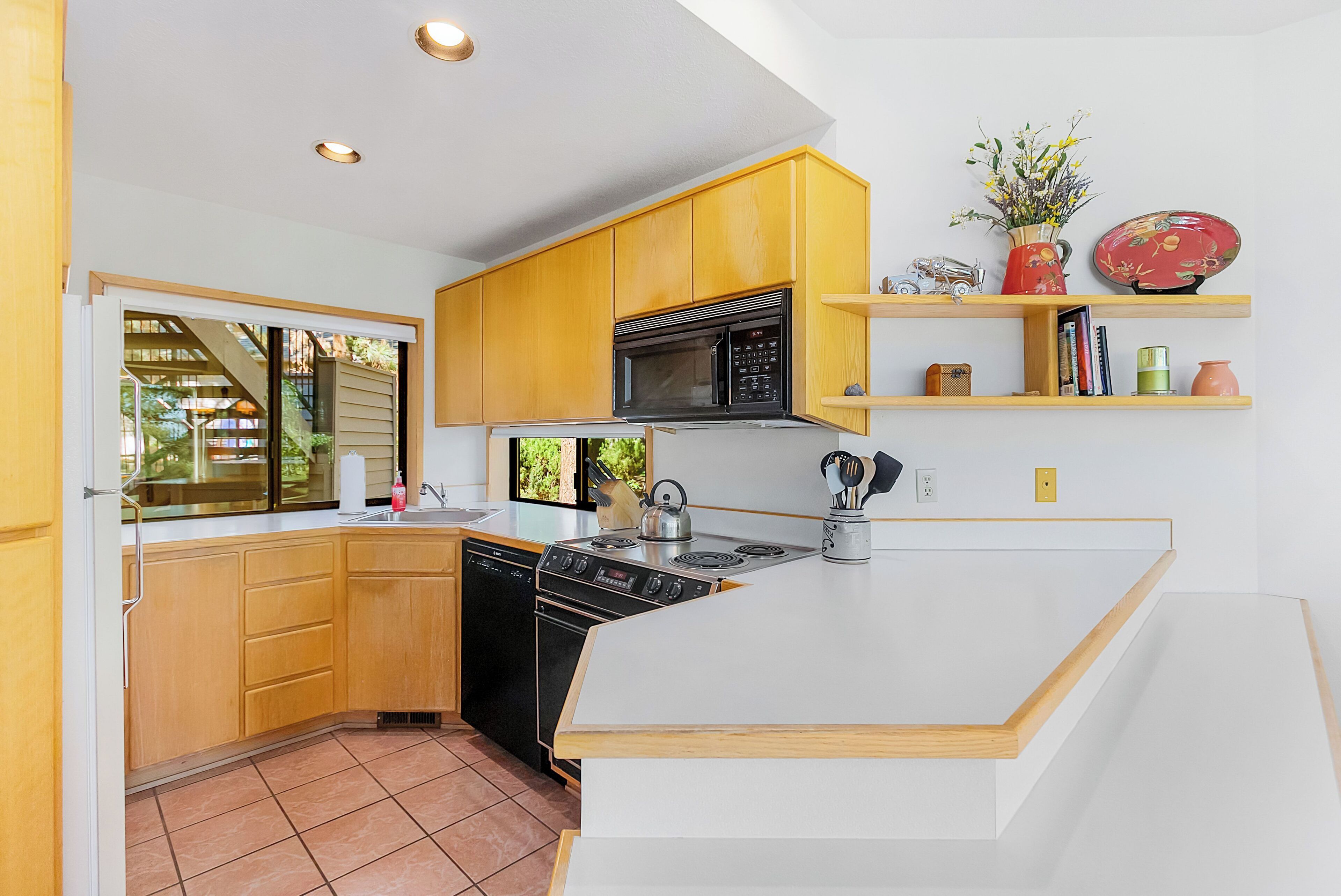 A kitchen with wooden cabinets, a microwave, a stove, and a sink. There are shelves with decorative items and a window with a view of greenery.