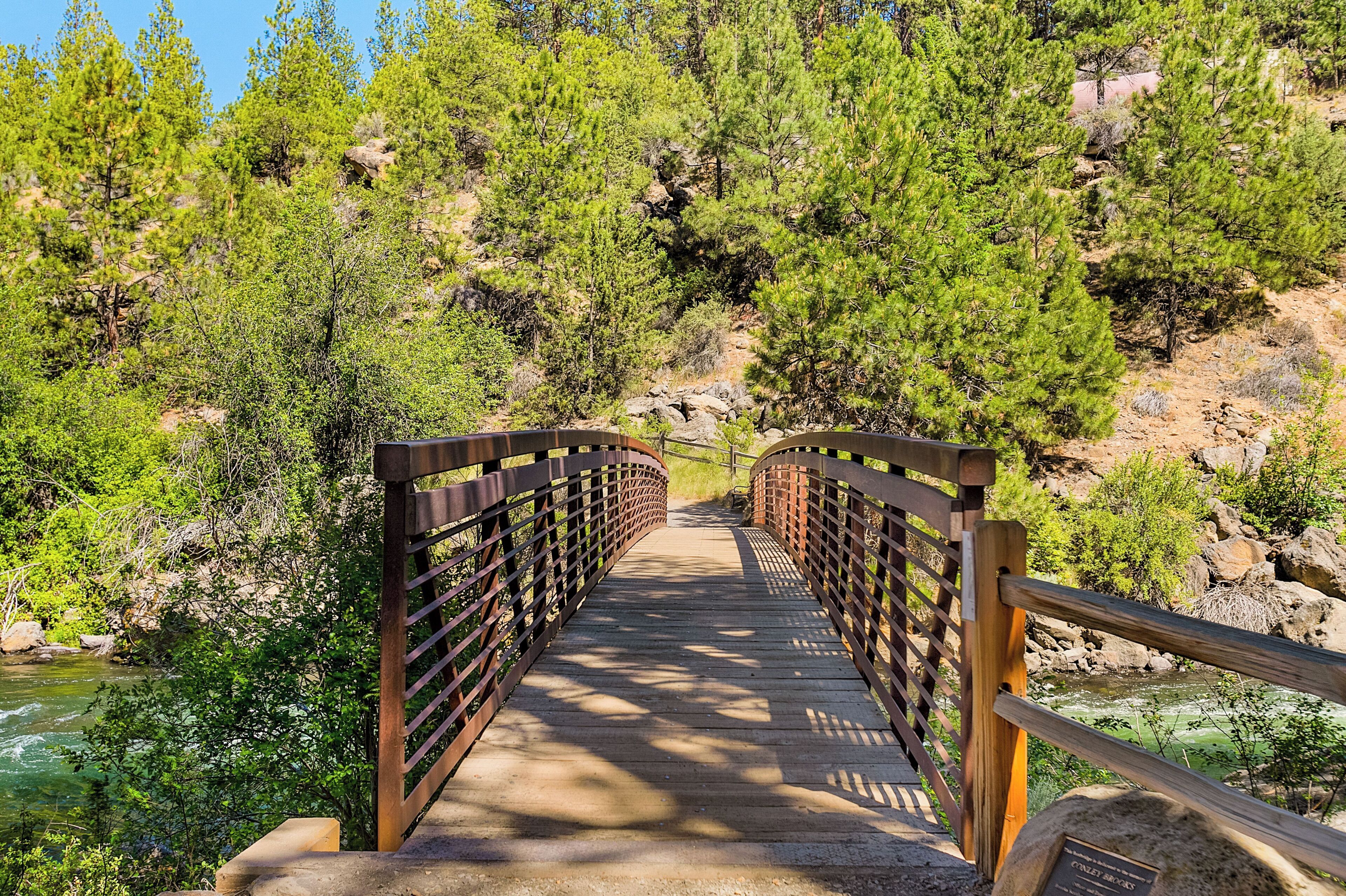 A wooden bridge with metal railings, surrounded by dense green trees and a river.