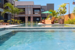 Un área de piscina moderna con agua cristalina, rodeada por una cerca de madera, y con un edificio de varios pisos con balcones y ventanas amplias.