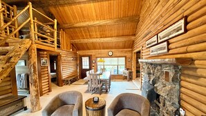 A log cabin interior with a dining area, two armchairs, a fireplace, and a wooden staircase. - Stanley House - Hillside Cabin with Panoramic Views of the Sawtooth Mountains (Stanley)