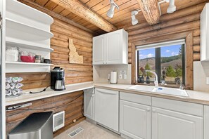 A kitchen with wooden walls, white cabinets, a coffee maker, and a window with a view of a mountainous landscape.