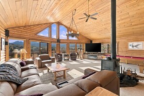 Un vaste espace de vie comprenant un grand canapé, une table basse et un téléviseur. De nombreuses fenêtres offrent une vue sur les montagnes.