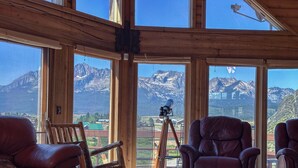 A room with large windows offering a mountain view, leather chairs, a telescope, and a wooden table. - Stanley House - Hillside Cabin with Panoramic Views of the Sawtooth Mountains (Stanley)