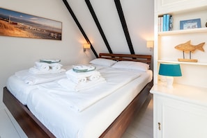A hotel room with a bed, towels, a painting, a lamp, and a bookshelf with books and a wooden fish decoration. - Ananda, App. 13 - Gemütliche 2-zimmer Og-ferienwohnung auf ca. 45 m² Wohnfläche, für 2 Personen (Wenningstedt-Braderup (Sylt))