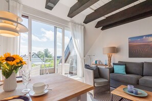 A modern living room with a sofa, a dining table with a vase of sunflowers, and a view of the outdoors. - Ananda, App. 13 - Gemütliche 2-zimmer Og-ferienwohnung auf ca. 45 m² Wohnfläche, für 2 Personen (Wenningstedt-Braderup (Sylt))