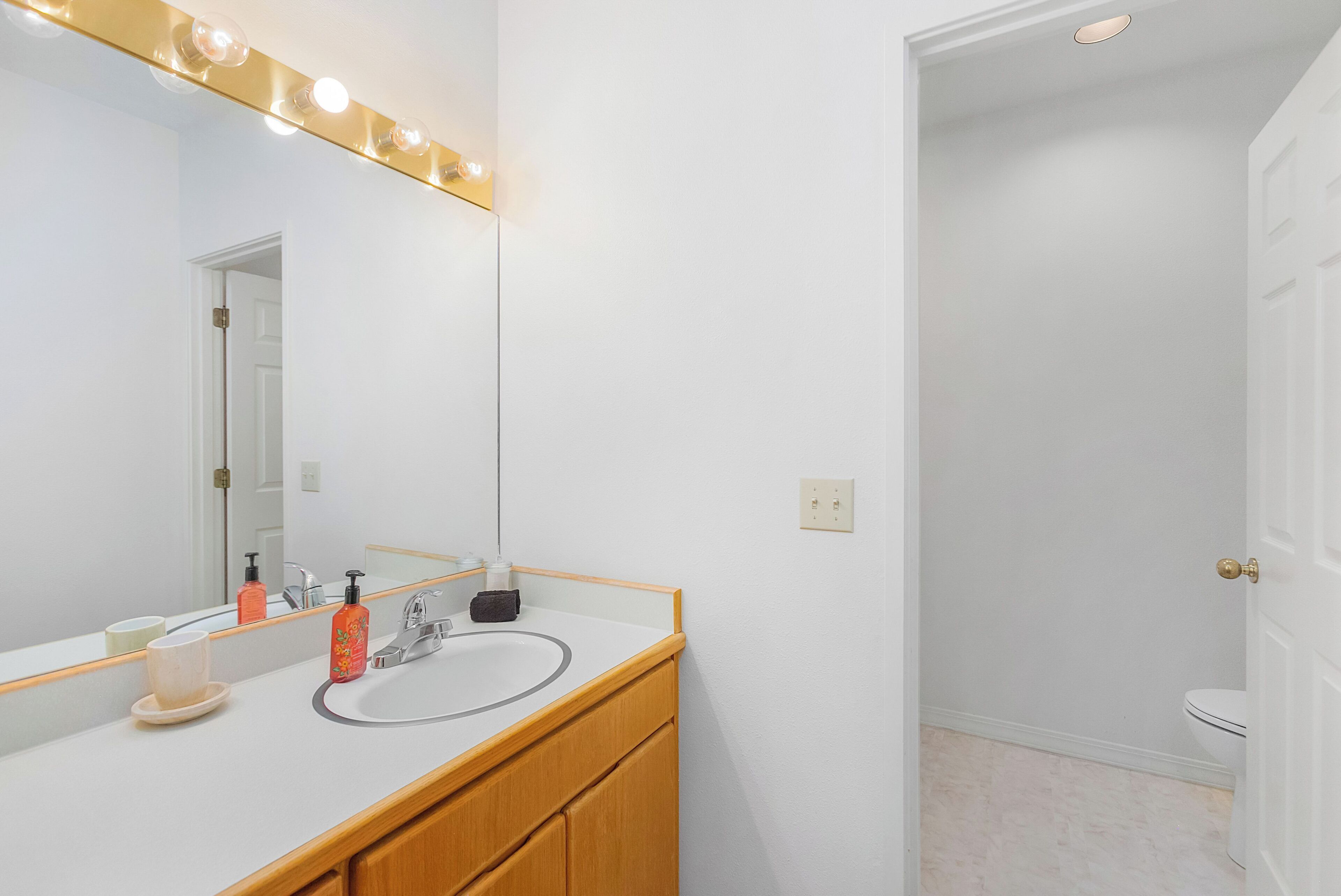 A bathroom with a white sink, a mirror with lights, and a door leading to another room.