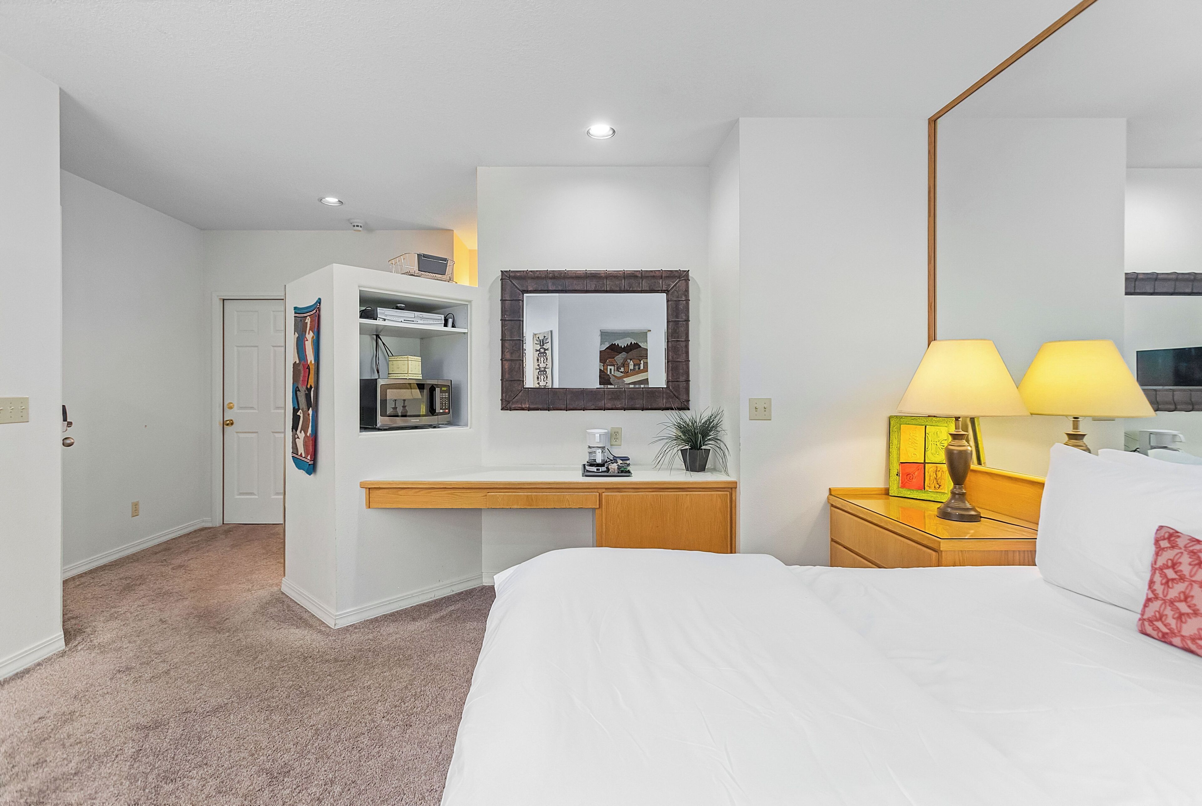 Een hotelkamer met een bed, bureau, stoel, nachtkastje, lamp, spiegel, een aan de muur bevestigde televisie en een deur.