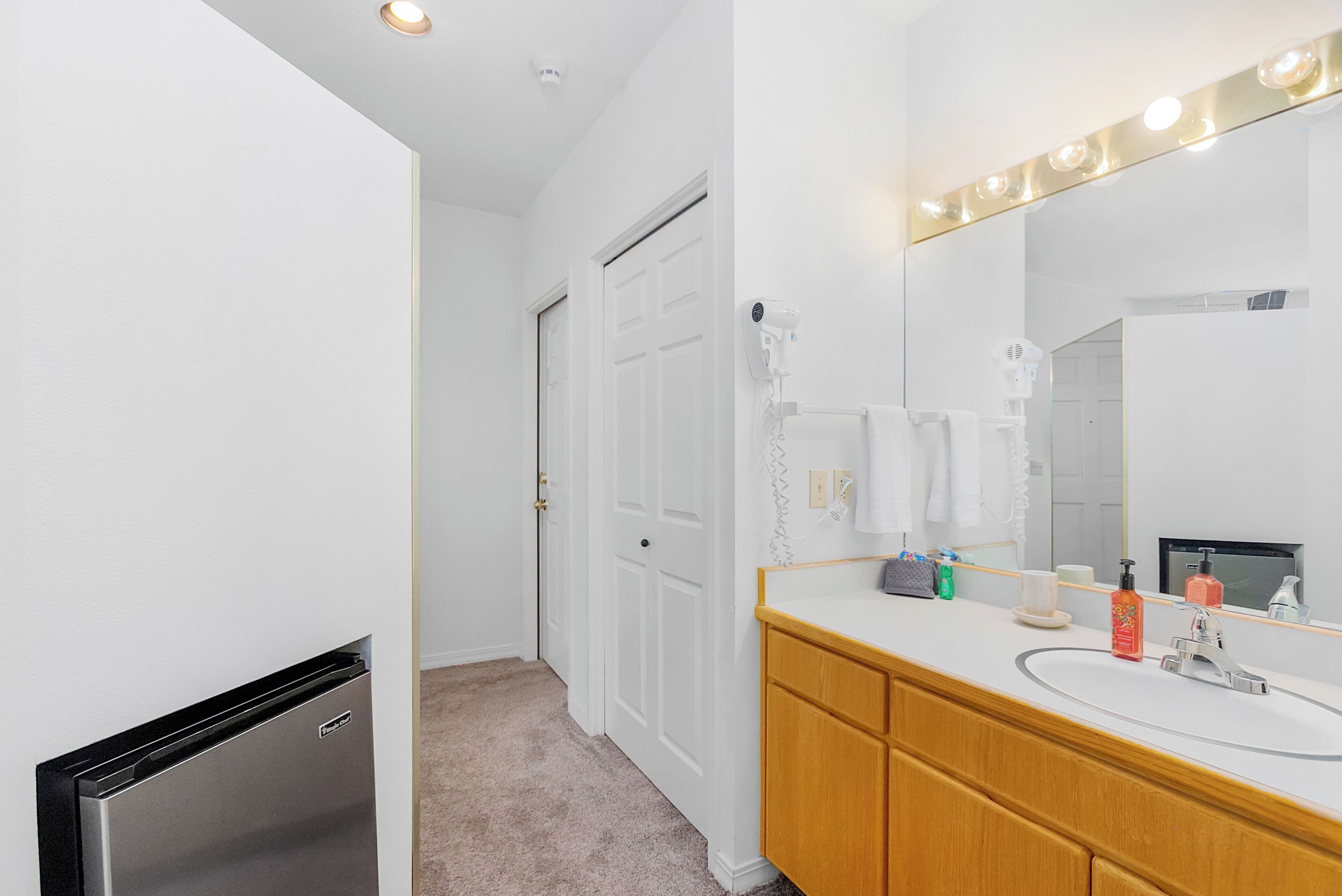 Une salle de bain moderne avec un meuble-lavabo blanc, un grand miroir avec éclairage intégré et une porte donnant sur une autre pièce.