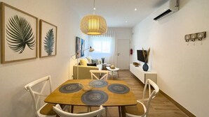A modern living room with a dining area, featuring a wooden dining table with placemats, white chairs, a hanging light fixture, and framed botanical prints on the wall.