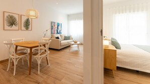 A modern living room with a wooden dining table, white chairs, a sofa, and a bed with a nightstand.