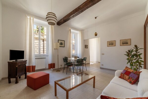 Un salón amplio con un sofá blanco, una mesa de centro de vidrio, un reposapiés rojo, una zona de comedor con una mesa redonda y sillas, y un televisor sobre un mueble de madera.