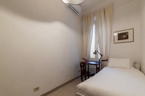 A bedroom with a bed, a desk, a chair, an air conditioner, a window with curtains, and a framed picture on the wall. - Panisperna 193 - FromHometoRome (Roma)