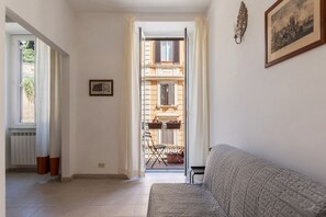 A room with a sofa, a framed picture, a window with curtains, and a view of a balcony with a chair and potted plants. - Panisperna 193 - FromHometoRome (Roma)