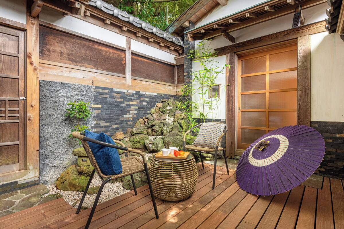 Un área de estar al aire libre de estilo japonés tradicional con esteras de tatami, un muro de piedra y una terraza de madera.