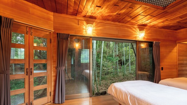 Une pièce en bois avec un lit, des portes-fenêtres coulissantes donnant sur une zone boisée et deux lampes fixées au mur.