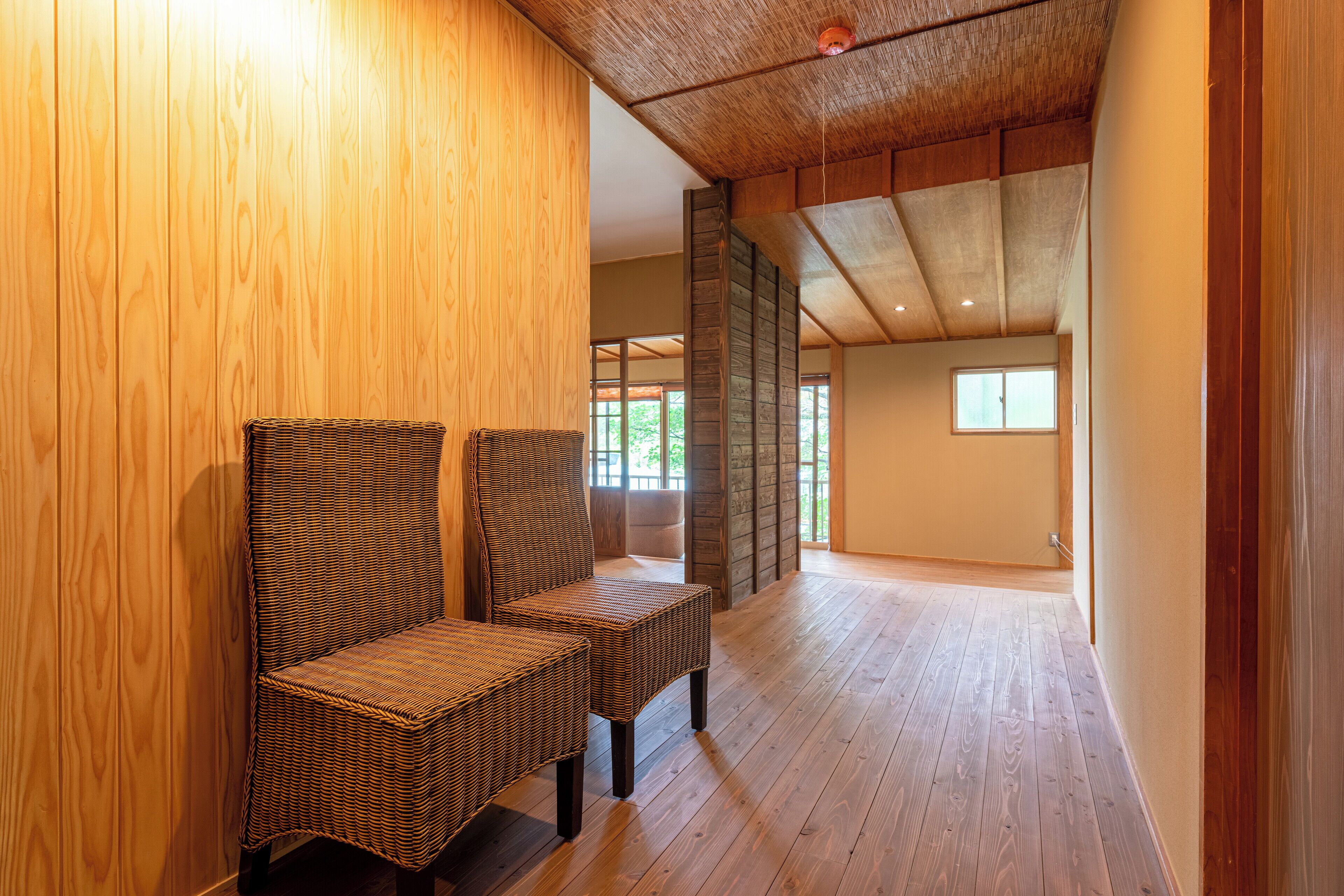 A spacious room with wooden flooring and a high ceiling. There are two wicker chairs and a large window providing natural light.