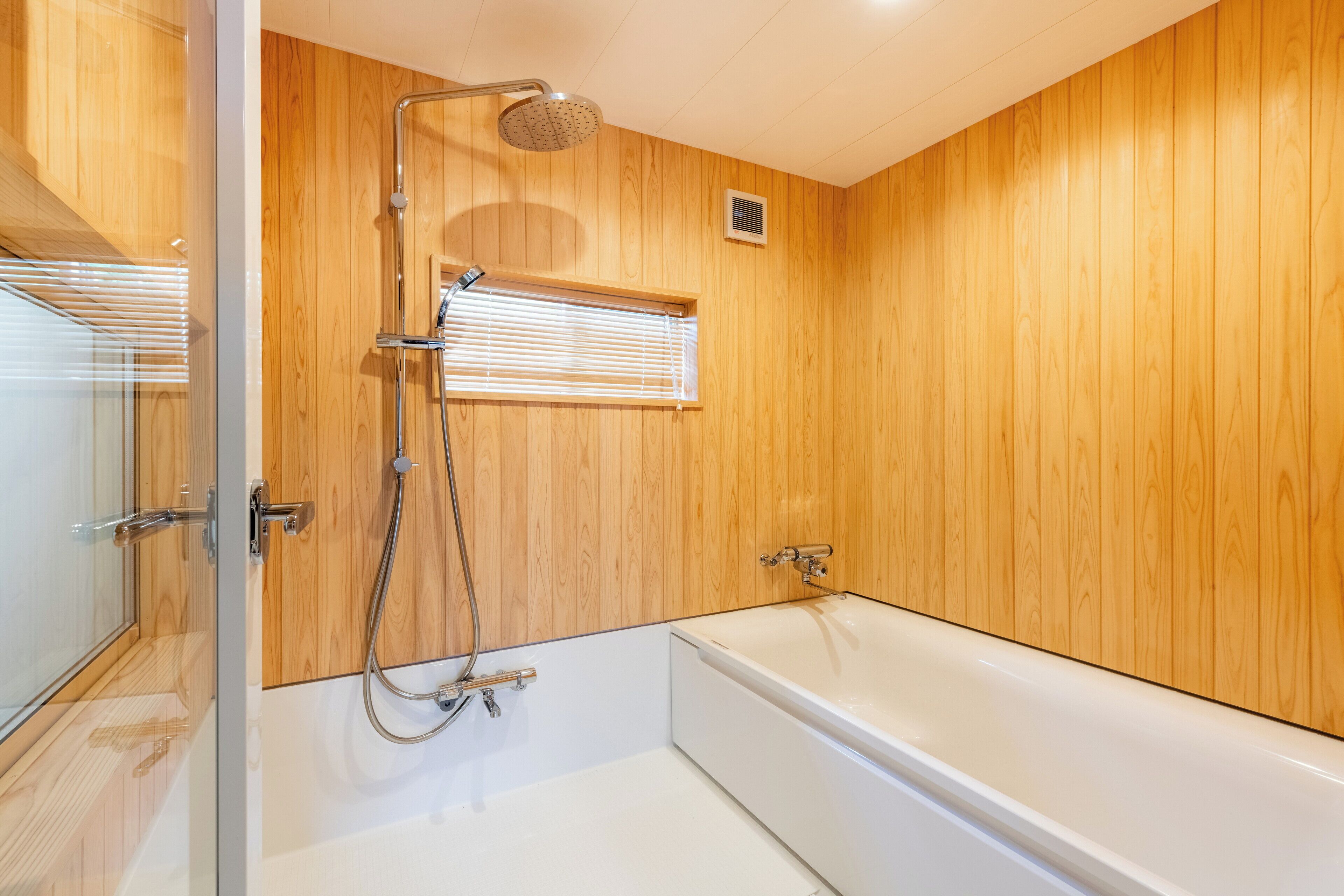 A modern bathroom with wooden paneling, a glass shower enclosure, a bathtub, and a window with blinds.