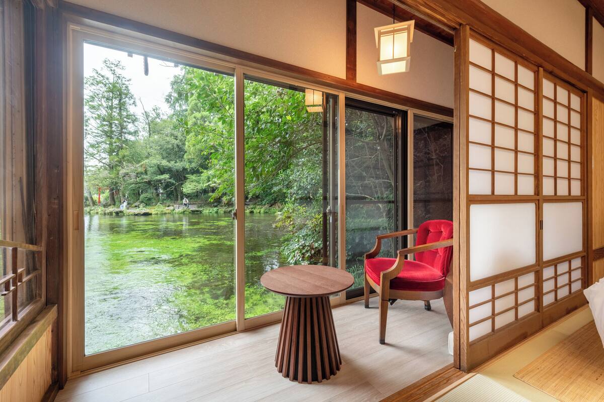 Una habitación con un amplio ventanal corredera que da a un estanque, una mesa de madera y una silla roja.