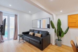 Una sala de estar moderna con un sofá de cuero negro, una mesita auxiliar pequeña, una planta grande en maceta y un balcón con puerta corredera.